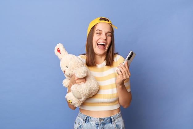 Indoor shot of happy joyful delighted teen girl wearing striped Tshirt and baseball cap holding cell phone and rabbit toy laughing happily being in good mood standing isolated over blue background