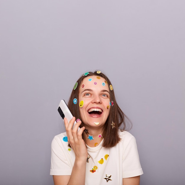 Indoor shot of happy joyful woman covered with funny stickers posing isolated over gray background talking phone and laughing copy space for promotion