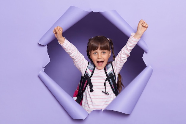 Indoor shot of happy little girl with pigtails wearing striped shirt and with backpack breaks through purple paper background raised arms yelling happily celebrating her victory rejoice