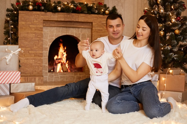 Indoor shot of happy parents posing near fireplace and Christmas tree with their infant daughter, positive father and mother sitting on floor on soft carpet with cute baby during New year time.