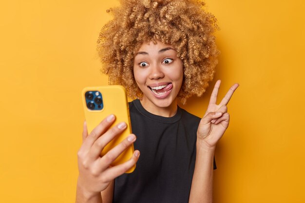 Indoor shot of happy positive curly haired woman sticks out tongue makes peace gesture takes selfie via mobile phone makes photo of herself wears casual black t shirt isolated over yellow background