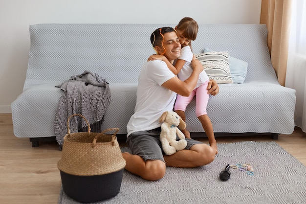 Indoor shot of happy positive young adult father spending time with his daughter while sitting on floor near sofa in living room, family hugging with love and gentle, playing together.