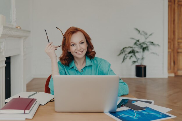 Indoor shot of happy redhead young European woman works on laptop computer communicates online sits in coworking space with paper documents holds spectacles wears blue shirt browses web