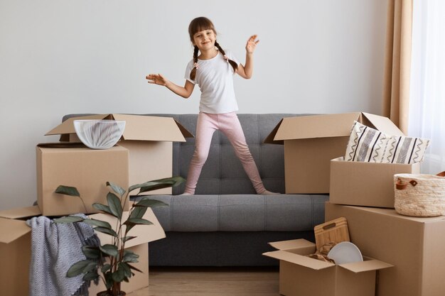 Indoor shot of happy smiling little girl wearing casual style attire standing on sofa and dancing being happy to move into a new apartment expressing positive emotions