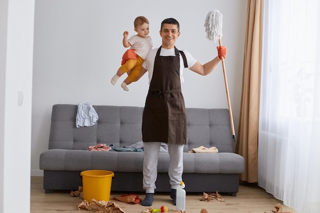 Indoor shot of happy strong man wearing casual attire and brown apron cleaning house with baby holding mop and raising arms showing his power looking at camera with optimism washing floor