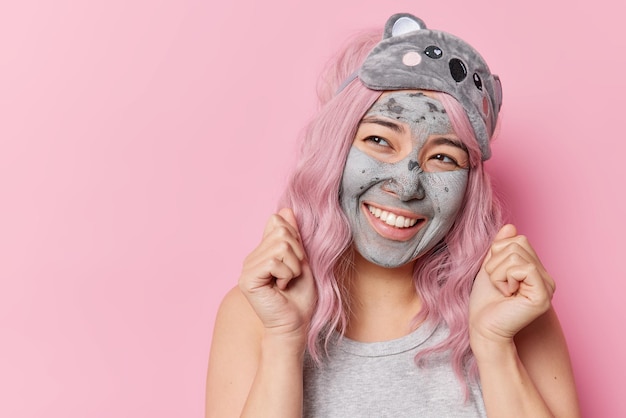 Photo indoor shot of happy woman clenches fists celebrates good news applies clay mask for skin rejuvenation has joyful dreamy expression wears sleepmask on forehead poses against pink studio wall