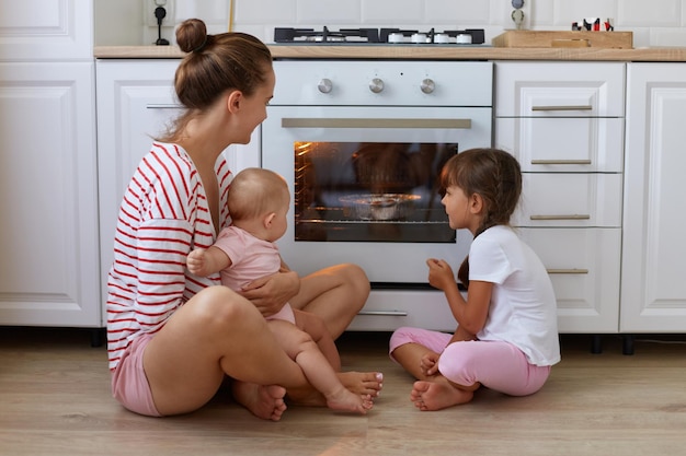 Photo indoor shot of happy woman with bun hairstyle wearing striped casual style shirt sitting on floor in kitchen with her children mother and her kids waiting for tasty dessert baking in oven