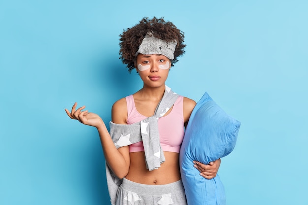 Indoor shot of hesitant puzzled woman raises arm looks with clueless expression at front dressed in nightwear holds soft pillow isolated over blue wall