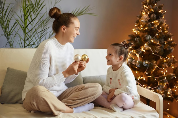 Indoor shot of joyful happy optimistic woman and her little daughter celebrating New year at home sitting on the sofa and having fun with Christmas balls expressing happiness being in festive mood