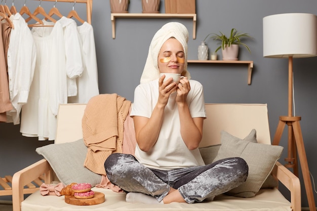 Indoor shot of joyful relxed woman wearing home clothes and with towel on head sitting on sofa with clothes hang on shelf on background holding cup of coffee and smelling aromat