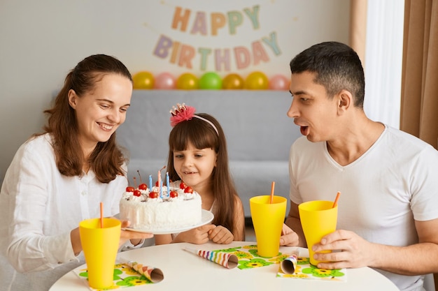 Indoor shot of little girl blowing out candles in her birthday with her family sitting at table together posing against balloons and holiday inscription on background