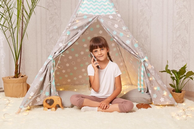 Indoor shot of little girl with pigtails wearing white t shirt playing in wigwam alone sitting on the floor and talking via with telephone with her friend looking at camera