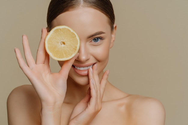 Indoor shot of lovely female model has fresh healthy skin well cared body covers eye with slice of orange poses bare shoulders