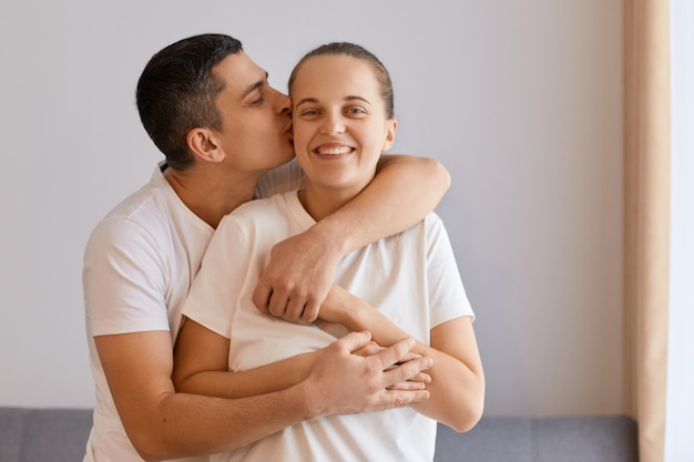 Photo indoor shot of loving husband cuddling wife from the back while standing in living room enjoying weekend celebrating anniversary expressing love and gentle smiling