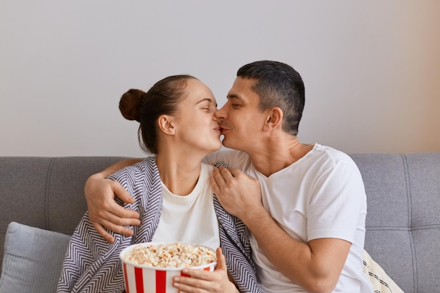 Indoor shot of loving romantic wife and husband sitting on sofa with pop-corn and remote control, watching romantic film, kissing while, expressing love and gentle.