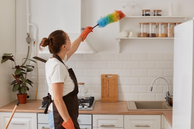 Inquadratura interna della cameriera che pulisce la polvere con ppduster in cucina casalinga con acconciatura da panino che fa le faccende domestiche pulendo la sua casa indossando maglietta bianca e grembiule