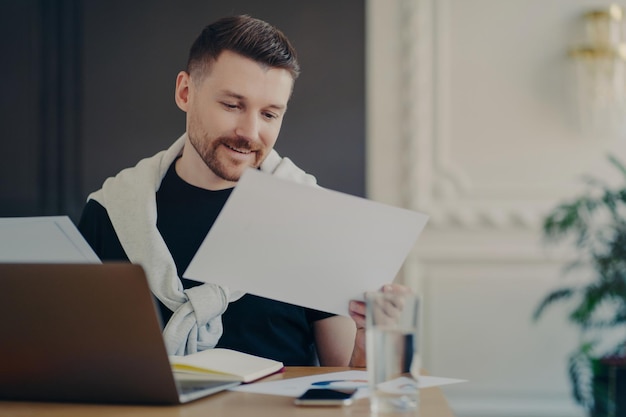 Indoor shot of male freelancer concentrated at papers prepares project or financial report poses at desktop with laptop computer works fron home blurred cozy background wears casual clothes