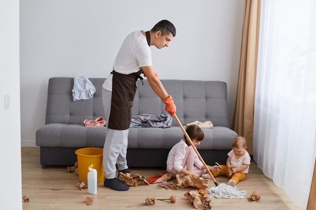 Снимок в помещении: мужчина в повседневной одежде и фартуке, моющий пол, позирует со своими дочерьми возле серого дивана, занимаясь домашними делами с сердитым выражением лица