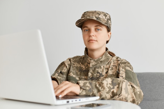 Indoor shot of military woman wearing camouflage uniform and\
cap sitting looking at camera with serious facial expression typing\
on keyboard working online on notebook