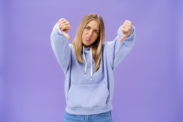 Indoor shot of moodie displeased and unimpressed cool modern female with blond hair and tanned skin tilting head sticking out tongue showing thumbs down in dissatisfaction posing against purple wall