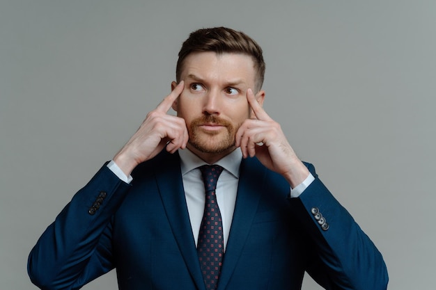 Indoor shot of motivated thoughtful bearded businessman keeps fingers on temples looks away gets ready for conference dressed in formal clothes isolated over grey background has hard working day