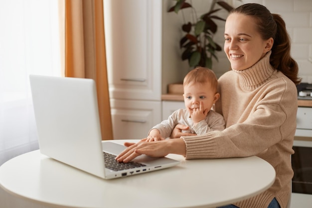 写真 キッチンに座って、娘と時間を過ごし、ビジネスパートナーとの会議を持っているラップトップでオンラインで作業しているベージュのセーターを着ている幸せなポジティブな愛らしい女性の屋内ショット