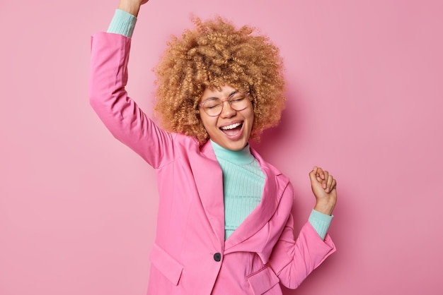 Фото Снимок в помещении счастливой оптимистичной женщины с вьющимися волосами трясет руками, заставляет триумфально танцевать, громко восклицает, носит очки, официальную куртку, изолированную на розовом фоне. успешная деловая женщина чувствует радость