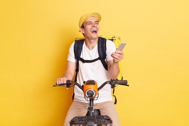 黄色の背景に分離された肯定的な感情を表現する笑いながら自転車に乗ってキャップとサーモ バッグを身に着けている携帯電話を保持している幸せな若い配達人の屋内ショット