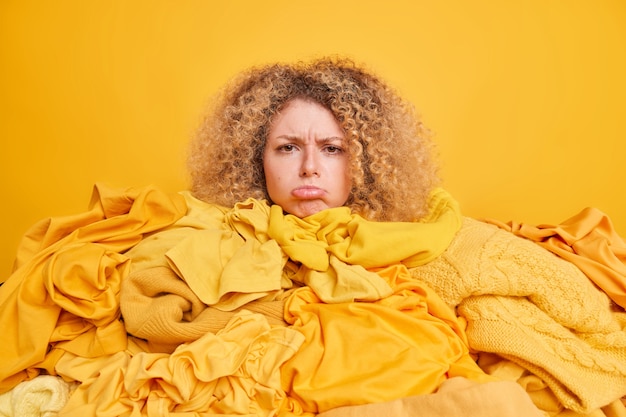 Фото Снимок в помещении: расстроенная кудрявая европейка в окружении белья, заваленного сумками для одежды, губы расстроили выражение лица, изолированное желтым