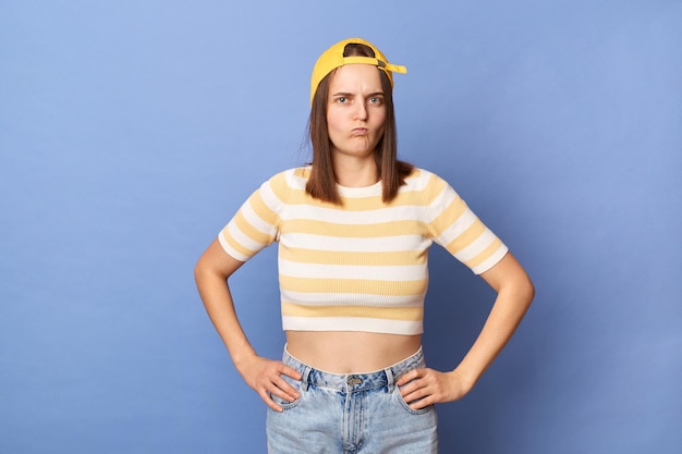 줄무늬 티셔츠와 파란색 배경 위에 격리된 야구 모자를 쓴 화가 난 10대 소녀의 실내 사진은 엉덩이에 손을 얹고 뺨을 불고 있습니다.