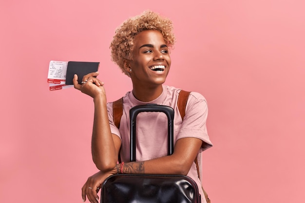 Indoor shot of optimistic Afro female tourist with luggage holds passport and tickets waits for flight ready to vacation enjoys travel receives visa for traveling abroad dressed in casual outfit