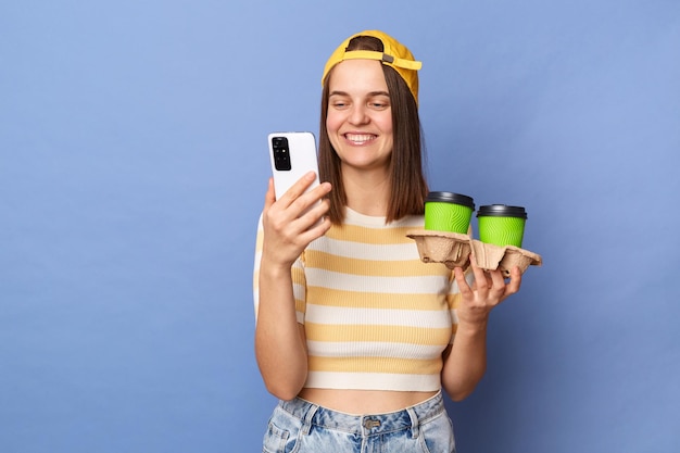 Indoor shot of optimistic charming teenager girl wearing striped Tshirt and baseball cap posing isolated over blue background holding double coffee to go and using mobile phone