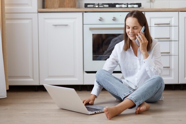 Indoor shot of optimistic dark haired woman wearing white t shirt and jeans sitting on floor in kitchen and talking phone while working online on portable computer.