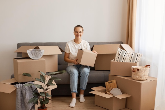 Indoor shot of optimistic good looking woman wearing white casual t-shirt and jeans, sitting on sofa, surrounded with boxes during relocating,holding package with personal pile, looks happy