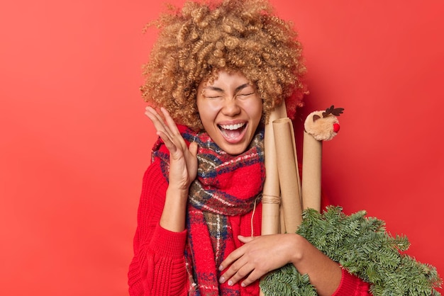 Photo indoor shot of overjoyed emotional curly haired woman exclaims loudly keeps hand raised has happy expression anticipates winter holidays poses against red background. yeah new year is coming