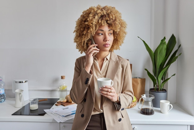 Indoor shot of pensive beautiful curly haired woman dressed\
elegantly has telephone conversation and drinks coffee poses at\
home kitchen talks with business partner thinks over future\
plans