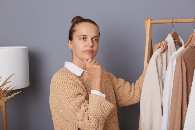 Indoor shot of pensive thoughtful young adult woman wearing beige knitted sweater standing near shelves with clothes choosing new attire thinking about the most trendy look