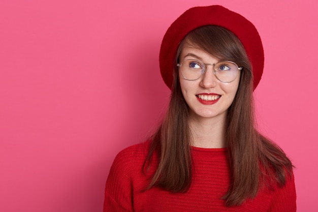 잠겨있는 젊은 여성의 실내 샷 빨간 스웨터, 베레모 및 둥근 안경, 흥미로운 또는 교활한 계획을 세 심하게 옆으로 보인다.