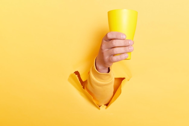 Indoor shot of person hand breaking through yellow paper and holding cup with water or other liquid showing mug with coffee or tea