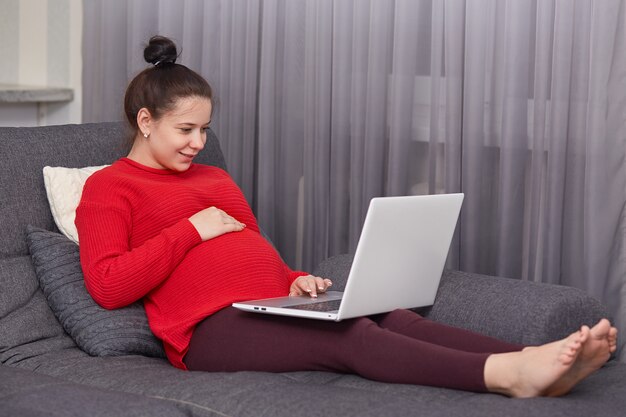 Indoor shot of pleased dark haired pregnant young woman enjoys domestic atmosphhere, keyborads information on laptop computer, sits barefoot on sofa, connected to wireless high speed internet