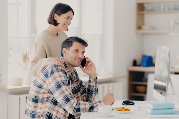 Indoor shot of pleased woman and man busy with discussing\
domestic expenses cheerful husband in checkered shirt makes\
telephone call caring wife touches shoulders focuse in monitor of\
computer
