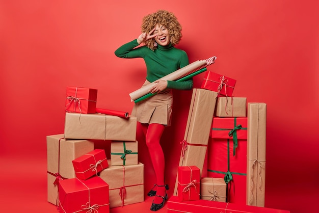 Indoor shot of positive curly haired woman smiles broadly makes\
peace gesture holds paper for wrapping gift boxes prepares presents\
isolated over red background winter holidays preparation