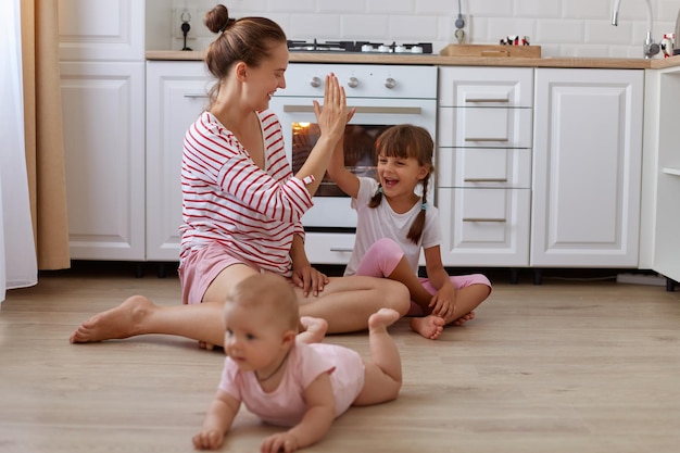 Indoor shot of positive optimistic woman wearing striped shirt sitting on floor in kitchen with her little daughters toddler kid crawling mommy giving five to her elder child