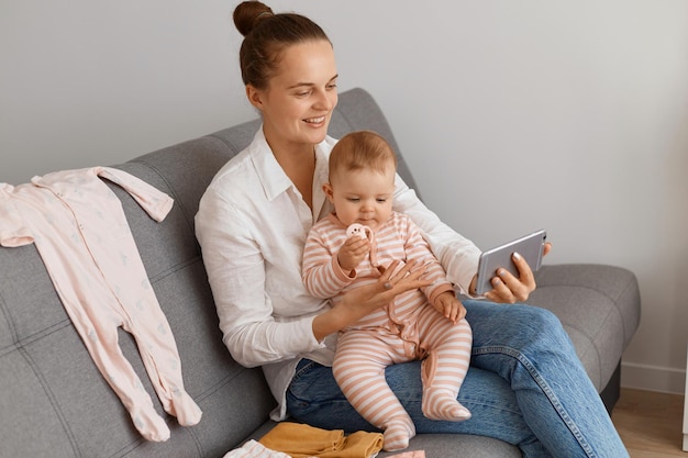 Indoor shot of positive satisfied female with bun hairstyle
wearing white shirt and jeans sitting on sofa with her infant baby
and holding smart phone in hands, having livestream in social
networks.