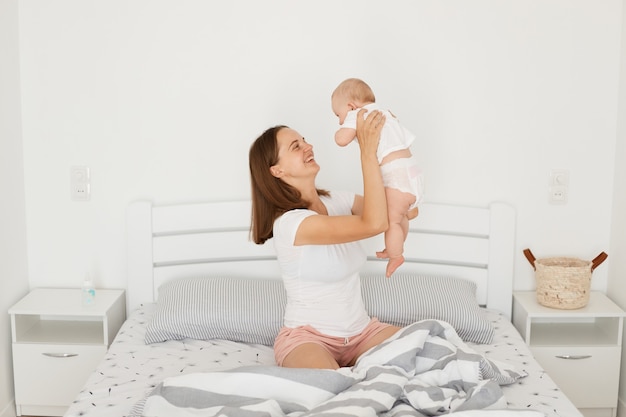 Indoor shot of positive smiling young mother playing with her infant baby while sitting on bed in light room, woman wearing white casual style t shirt and pink shorts, happy motherhood.