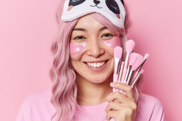 Indoor shot of positivev young asian woman smiles broadly applies hydrogel patches under eyes holds cosmetic brushes for applying makeup wears sleepmask on blindfold isolated over pink background