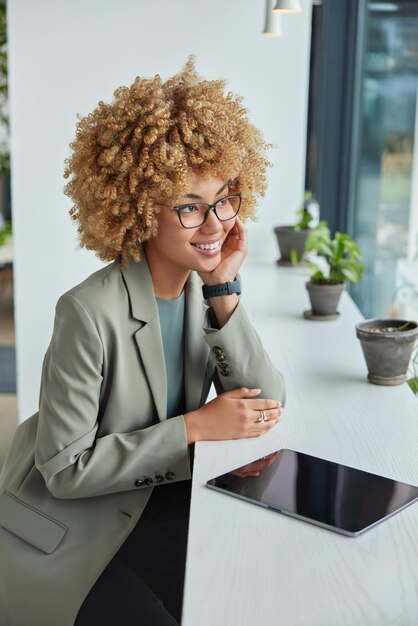 Indoor shot of positve businesswoman dressed formally has\
dreamy cheerful expression poses at windowsill with digital tablet\
on it browses news on digital device admires view through\
window