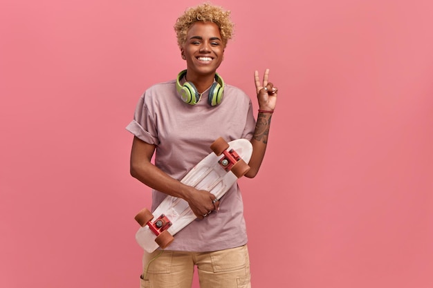 Indoor shot of pretty African American woman with glad expression, raises hand and makes peace sign, wears lilac t shirt, has headphones, holds skateboard, enjoys favourite hobby, over pink background