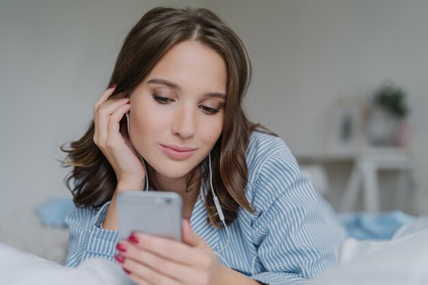 Indoor shot of pretty dark haired woman enjoys music in playlist lies on comfortabled bed at home has day off watches films online connected to wireless internet enjoys cozy atmosphere