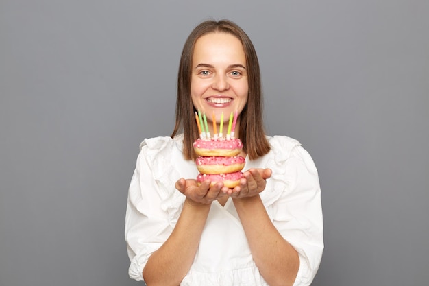 Indoor shot of pretty friendly pleased woman with brown hair wearing white blouse looking at camera with happiness holding donut with candles isolated over gray background